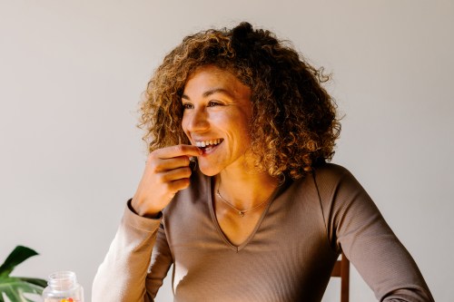 close up of person with brown curly hair in a light brown long sleeve shirt taking a probiotic supplement at their kitchen table to help the gut health and anxiety connection