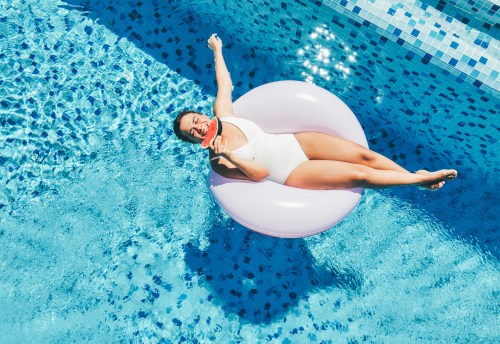 Woman relaxing on an inflatable ring and eating juicy watermelon in outdoor swimming pool on sunny day. Aerial view.