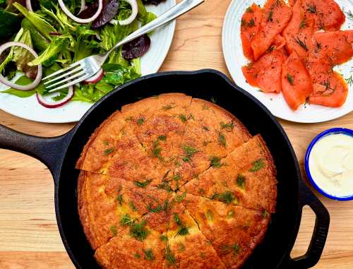 a chickpea pancake in a black cast iron pan on a wood cutting board surrounded by salad plates and smoked salmon