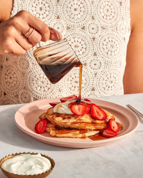 a woman pours maple syrup onto a stack of pancakes covered in strawberries