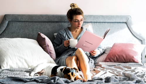 Pretty young woman sitting on her bed with her dog pet and reading a book while holding an ember mug.