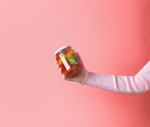 Female hand with light pink sweatshirt holding glass jar with various preserved food on pink background.