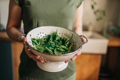 Girl in leggings and green shirt holding colander of washed spinach with hands visible, top view.