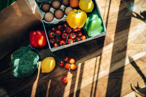 Flat lay of a home delivery cardboard box filled with multi-coloured fresh organic fruits, vegetables and groceries on wooden background against sunlight.