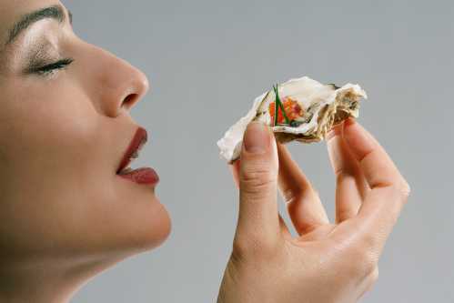 a woman wearing red lipstick holds up an oyster to her mouth