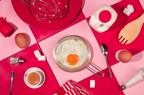 Pink and red tiled kitchen table, with various kitchen utensils that look similar to the Paris Hilton Walmart collection and ingredients. In the center there's a fried egg in a small frying pan.