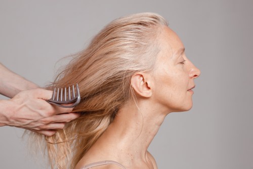 An unrecognizable male hairdresser combs the dried hair of middle aged blond woman sitting sideways