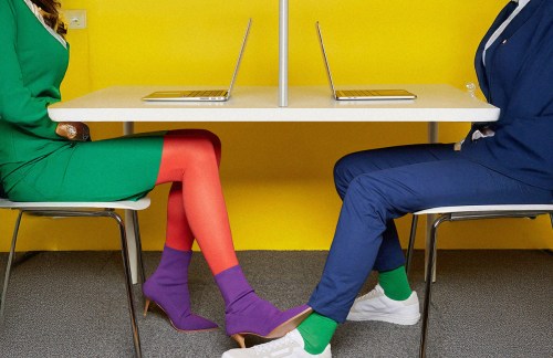 Two people's legs touch under a table as they work on their laptops.