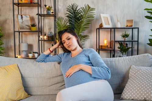 pregnant person in blue shirt and light grey pants sitting on couch holding stomach and feeling sad in the living room