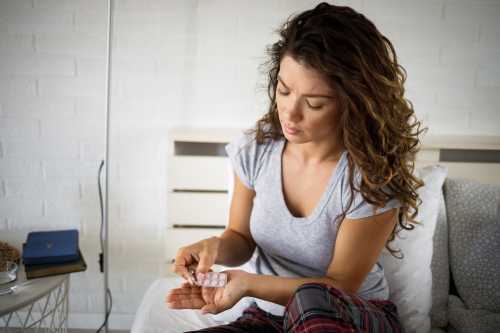 person with brown curly hair in a grey t-shirt and plaid pajama pants sitting on their bed and holding a metal medicine packet of antihistamines for PMDD