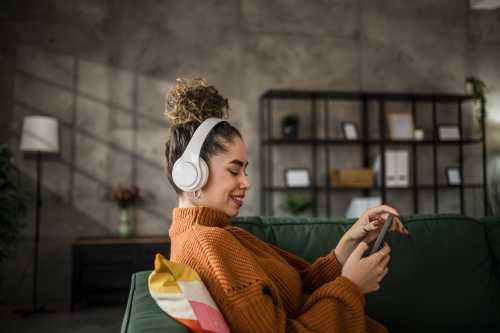 young woman in orange sweater listening to music on wireless headphones over a mobile app while sitting on a comfortable green sofa