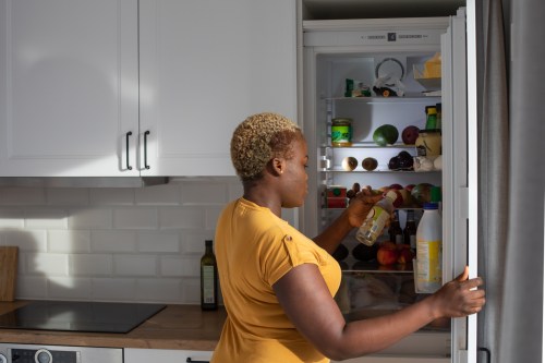 woman in yellow shirt opening up fridge in kitchen and checking the expiration date on a bottle