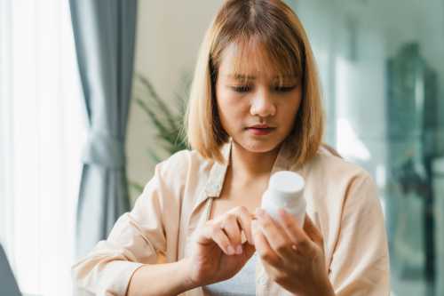 Close up image of a person in a pink button down shirt with a short blonde haircut looking at a bottle of probiotics while sitting at home, wondering can you take too many probiotics