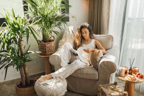 Woman at home reading book while sitting ona. beige chair and drinking coffee with candles lit on the table.