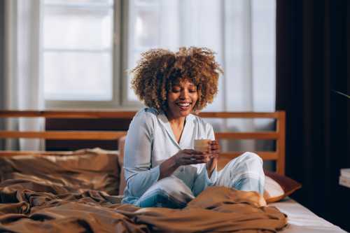 person with brown curly afro wearing blue cotton pajamas sitting cross-legged on their bed in the morning while drinking golden milk, learning how to stop feeling tired in winter