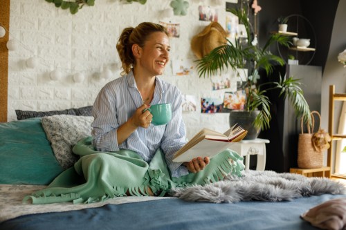 Cheerful woman enjoying morning coffee in bed while reading a book and using a mint green blanket