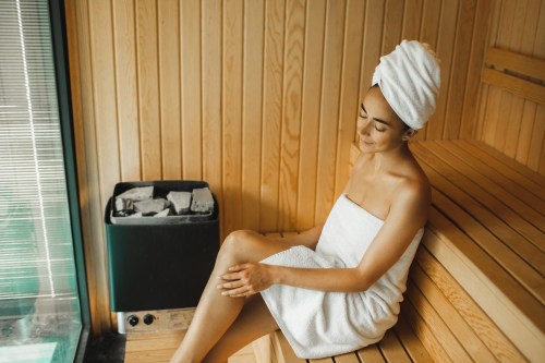 close up of person with tan skin wearing a towel around their head and one around their body while sitting inside a wooden sauna next to a coal stove, hoping to reap sauna therapy health benefits