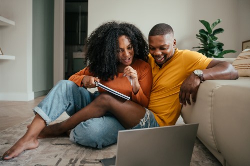 Couple wearing orange shirts and denim jeans sitting on the floor of their living room and taking notes on a notepad while looking at a laptop with free sex worksheets