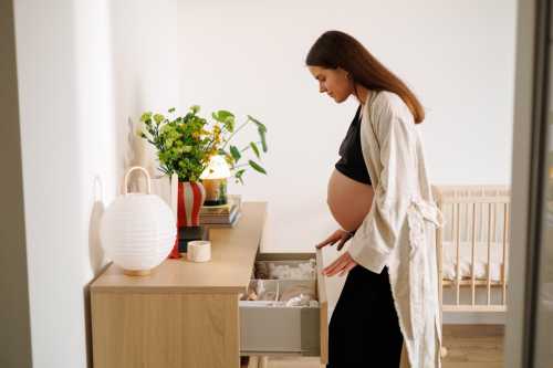 pregnant person with long brown hair in a black tank top and pants and flowy tan overshirt looking at their baby clothes and awaiting birth, to show concept of superfetation pregnancy