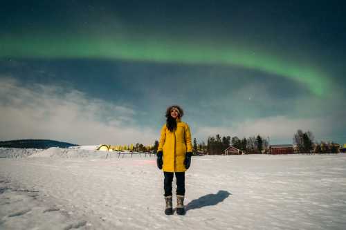 A woman is a yellow coat looks up at auroras in a snowy landscape.