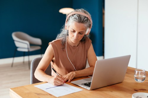 close up of person with long curly grey hair wearing pink headphones while sitting at their laptop taking notes on a notepad at their kitchen table, learning things to do in 50s to reduce dementia risk