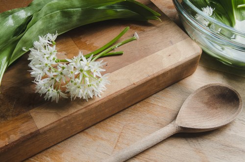 close up of wild garlic flower and leaves on a wooden table, wooden cutting board, and next to a wooden spoon and glass bowl because garlic can help a cold