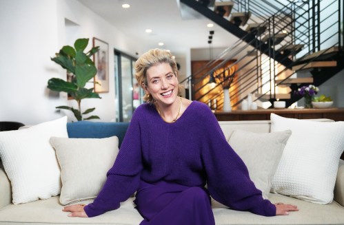 Actress Kate Walsh wearing a purple sweater and pants while smiling at the camera and sitting on her couch in her home