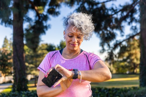 close up of person with grey curly hair wearing a pink short sleeve shirt standing outside while holding their phone and looking at their fitness tracker, getting ready for a morning walk while biohacking for longevity