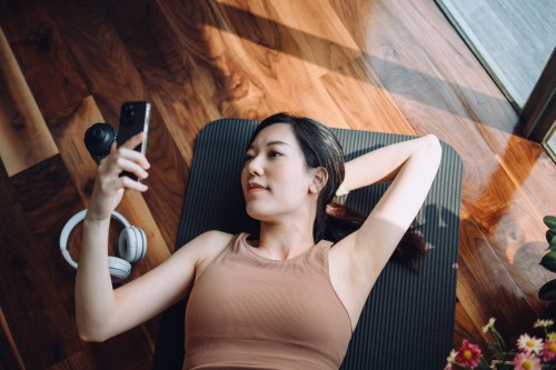 High angle shot of fitness young Asian sports woman relaxing after working out, lying on yoga mat and using her smartphone against sunlight in the morning, reusable water bottle and headphones by her side.