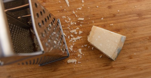 Grating Parmigiano-Reggiano. Grater partially in field of view. Shreds and piece of cheese on wooden chopping board. High point of view.