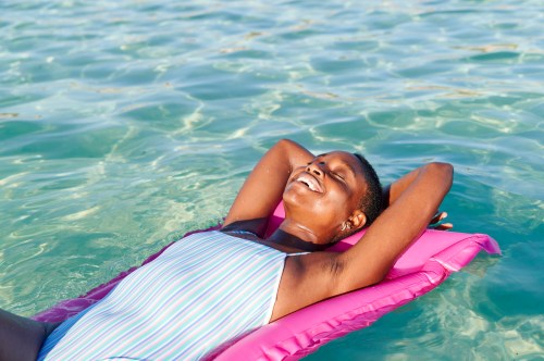 Smiling young African woman in a one piece swimsuit lying with her eyes closed on an inflatable float on the ocean