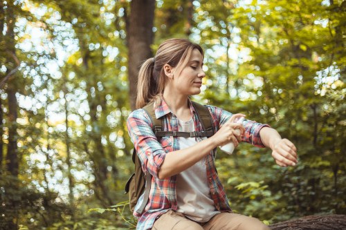 person sitting in the middle of the woods wearing a plaid flannel shirt and hiking backpack spraying bug repellent on their arm to avoid alpha-gal syndrome tick bites