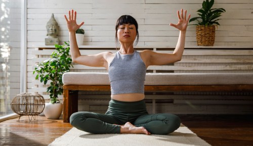 Woman doing arm exercise without weights while sitting on mat at home.