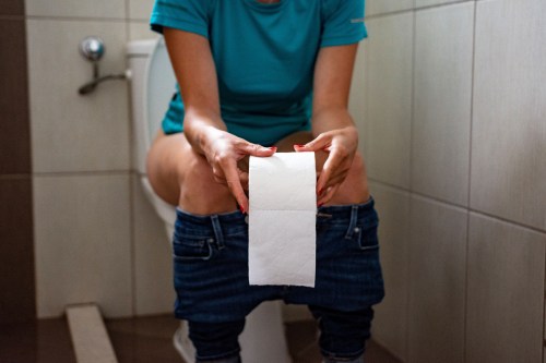 close up of person wearing a blue shirt and blue jeans sitting on the toilet while holding a roll of toilet paper and dealing with black poop