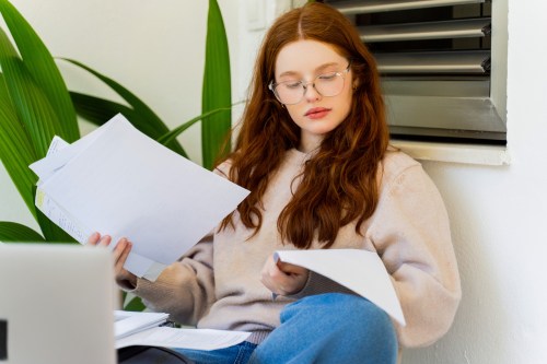 person with long red hair in a tan sweater and blue jeans sitting at home reading papers while wearing reading glasses, wondering how do you know if you need reading glasses