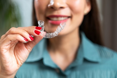 Close up of person in green collared shirt with bright red nail polish smiling while holding their clear dental aligner, learning how to clean a retainer for oral health