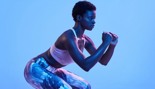 Side view of strong African American female in sportswear squatting during fitness workout under blue light