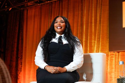 photo of singer Lizzo in a black dress and white long sleeve button down shirt and tie sitting and smiling for the camera against an orange velvet curtain background