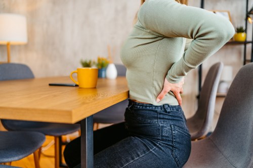 close up of person in jeans and a light green long sleeve shirt holding their lower back in pain while sitting at their kitchen table