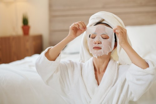 woman doing a morning shed routine while taking off a white sheet mask from face and wearing a white robe
