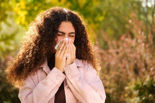 close up of person with long curly brown hair blowing their nose into a tissue while standing outside near trees, hoping to avoid the high pollen counts