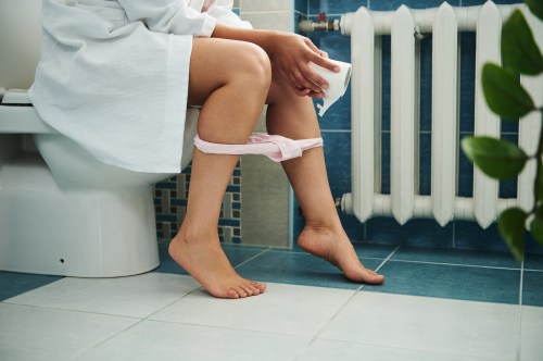 close up of person sitting on the toilet in their white bathrobe while holding a role of toilet paper, hoping that they don't have yellow poop