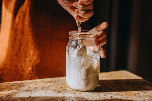 A hand is stirring a sourdough starter in a glass jar, on a kitchen countertop. The light casts a warm glow, highlighting the ingredients inside the jar.