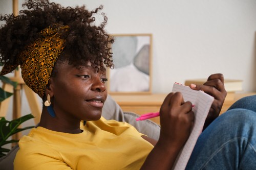close up of a person with black curly hair in a hair wrap wearing a yellow shirt and denim jeans writing in a journal and wondering why journaling makes me feel worse