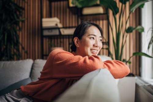 person in an orange sweater with their black hair pulled into a bun sitting on their couch at home in their living room while looking at the window, learning how to overcome burnout based on personality type