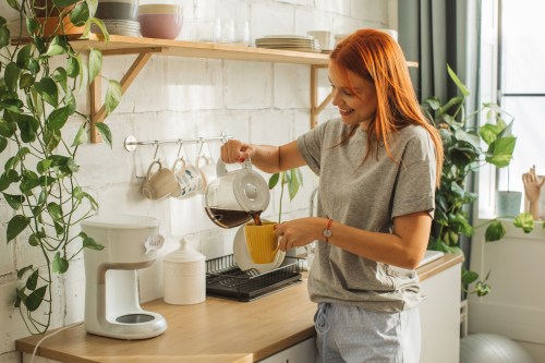 Woman in a sunlit room making coffee while checking smart phone.