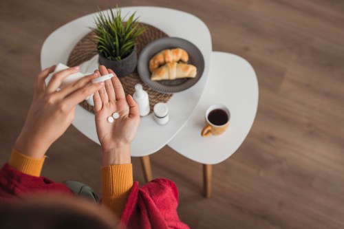 Aerial view on female hands taking pills from a medicine container above a coffee table with a mug of coffee and croissants