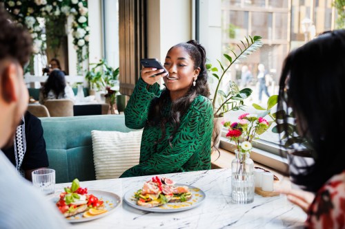 A delighted woman captures a photo of her colorful meal with a smartphone, sharing the vibrant dining experience at a beautifully decorated cafe with friends.
