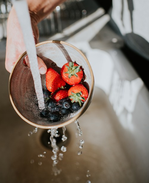 Fresh ripe strawberries being rinsed in a ceramic drainer under a running tap.