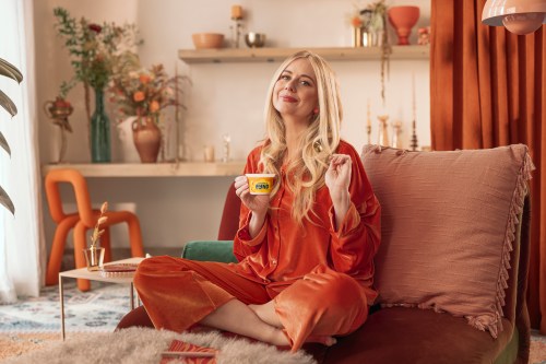 a blonde woman lounges on a red couch wearing red pajamas and holding a cup of yogurt.